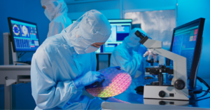 Cleanroom technician inspecting a silicon wafer beside a microscope and monitoring equipment