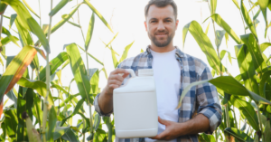 Farmer smiling and holding a white pesticide bottle in a corn field, ensuring crop protection and healthy growth.