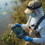 U.S. public health article about mosquito abatement and larval surveillance. Show a mosquito control technician in PPE standing at the edge of a marsh/wetland, performing larval monitoring with a dipper and sample tray while referencing a rugged tablet with a GIS map interface.